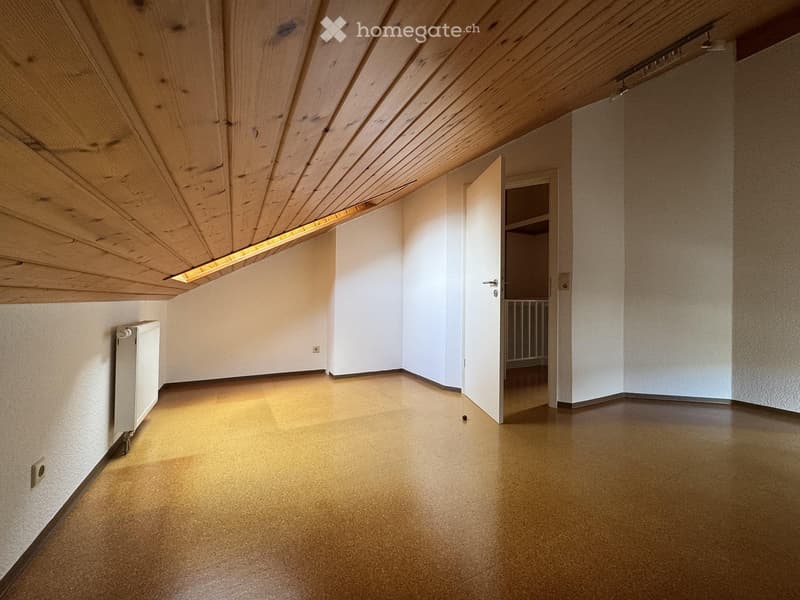 Empty room with wooden slatted ceiling, light-colored walls, and a smooth, light brown floor. A small white radiator is mounted on the left wall, and an open doorway leads to another room with a visible shelf unit.