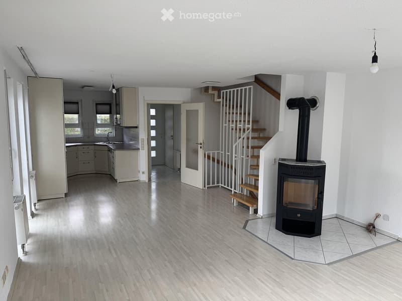 Open-plan living and kitchen area with light wood flooring, modern kitchen cabinets, a sink, a staircase leading to an upper level, and a black wood-burning stove.