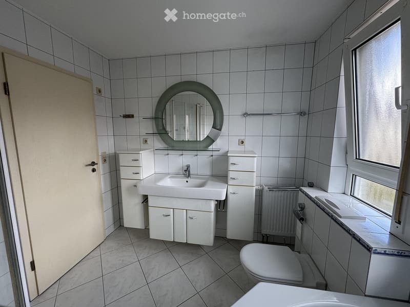 Modern bathroom with white tiles, a sink, a toilet, a round mirror, and white cabinets.