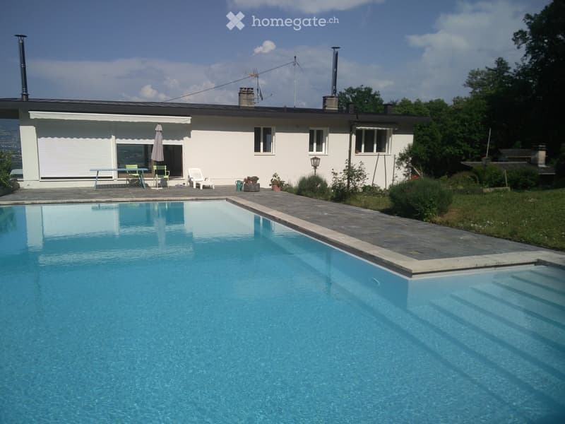 A modern house with a large swimming pool in the foreground, surrounded by a paved patio area with outdoor furniture and plants.