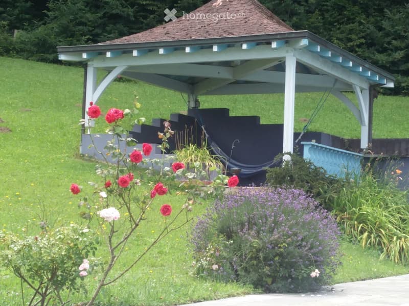 A gazebo with a wooden frame and a tiled roof is situated on a grassy lawn surrounded by colorful flowers and shrubs.
