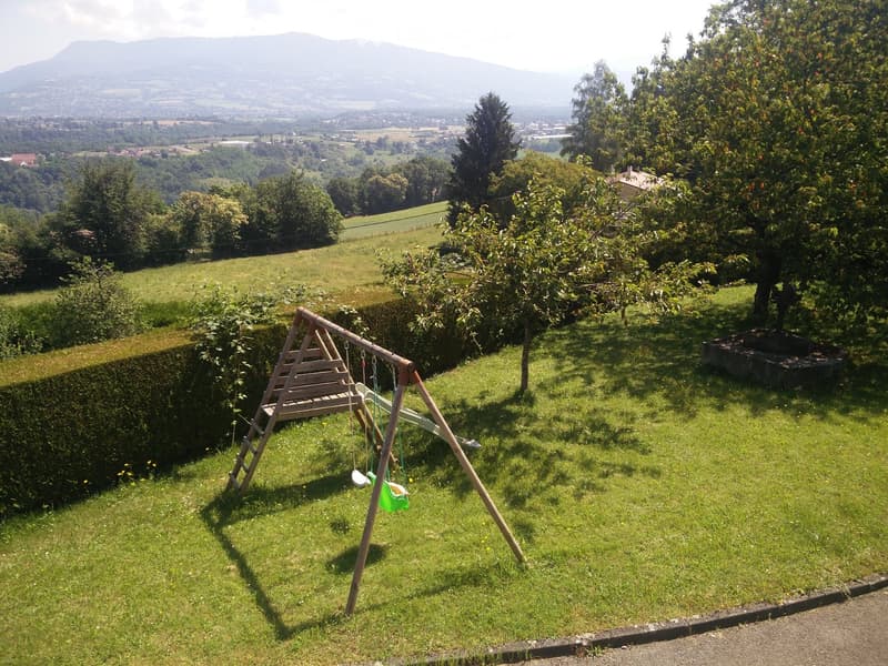 A grassy garden with a wooden swing set, a small slide, and a swing seat, surrounded by trees and a hedge, with a scenic mountain view in the background.