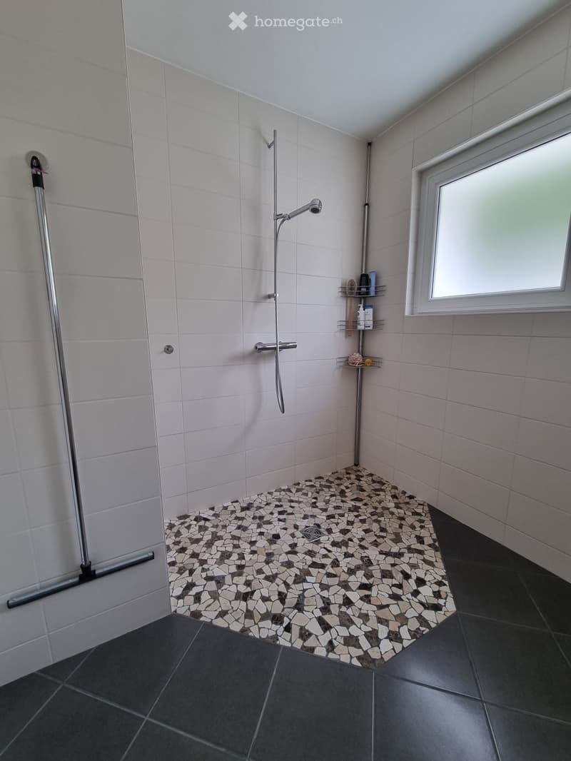 Modern shower area with white tiled walls, mosaic tile floor, chrome showerhead, and small shelf holding toiletries.