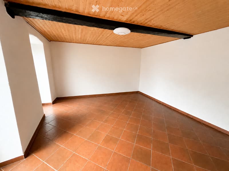 Empty room with white walls, terracotta tiled floor, and a wooden slanted ceiling with exposed beams. A single round light fixture is mounted on the ceiling.