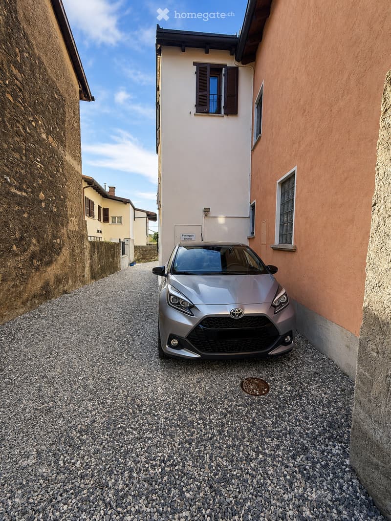 A narrow alleyway with a parked car, featuring a two-story building with white walls and brown shutters, surrounded by stone walls and a gravel surface.