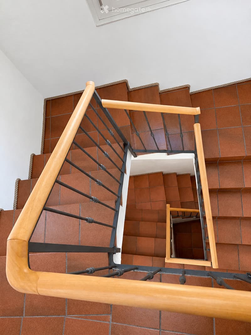 Spiral staircase with wooden handrails and metal balusters, featuring red brick walls and white ceiling.