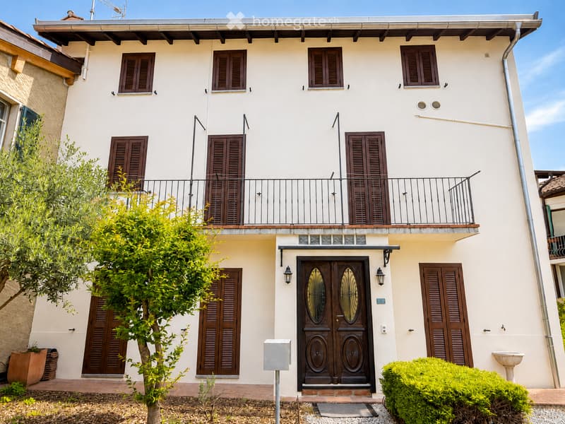 Two-story house with light beige walls, dark brown shutters, and a balcony on the upper floor. Features a large front door with decorative glass panels, a small tree, and a well-maintained garden area.