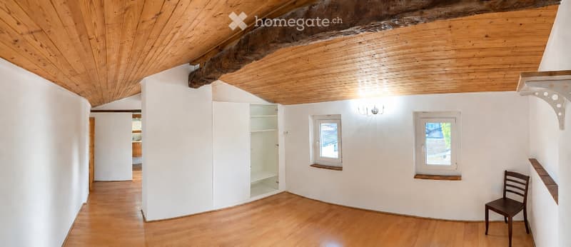 Empty room with wooden flooring, white walls, exposed wooden ceiling beams, and two windows. A single wooden chair is placed near the right wall.