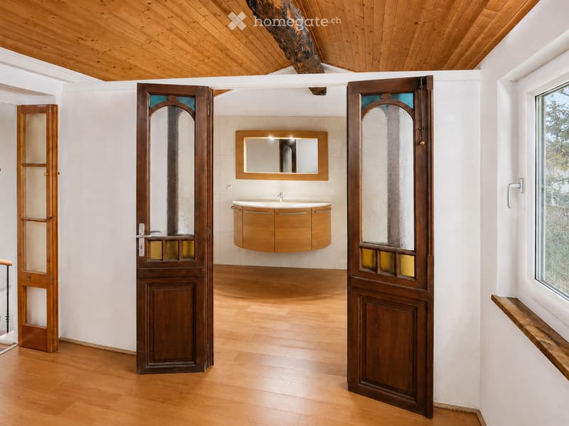 Modern bathroom with wooden flooring, white walls, a floating wooden sink cabinet, and a large mirror above it. The room features wooden doors with decorative glass panels and a window with a wooden sill.