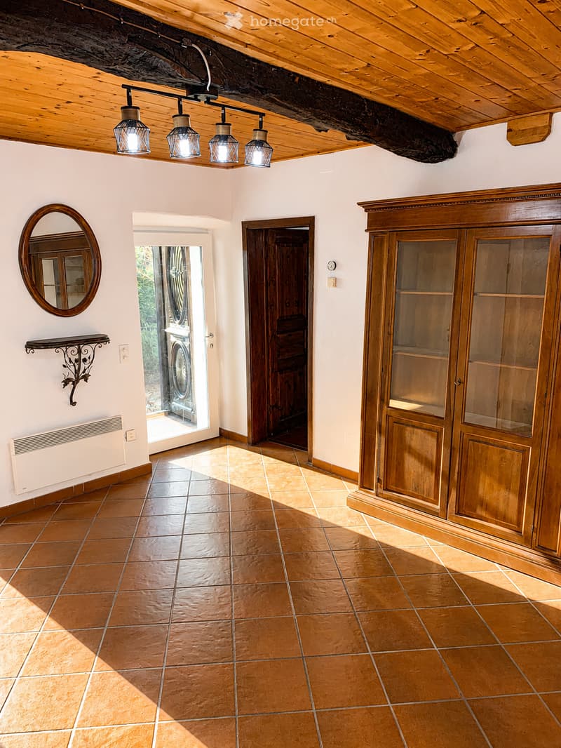Empty room with exposed wooden beams, terracotta tiles, a large wooden cabinet, a round mirror, and a decorative shelf. Natural light streams in from an open doorway.