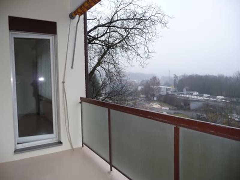 Balcony with glass railing, overlooking a cityscape with buildings and trees, glass door leading inside.