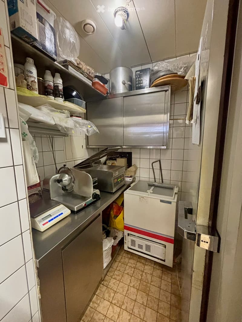 Commercial kitchen with stainless steel appliances, including a refrigerator, oven, and scale, along with shelves holding containers and plastic bags.