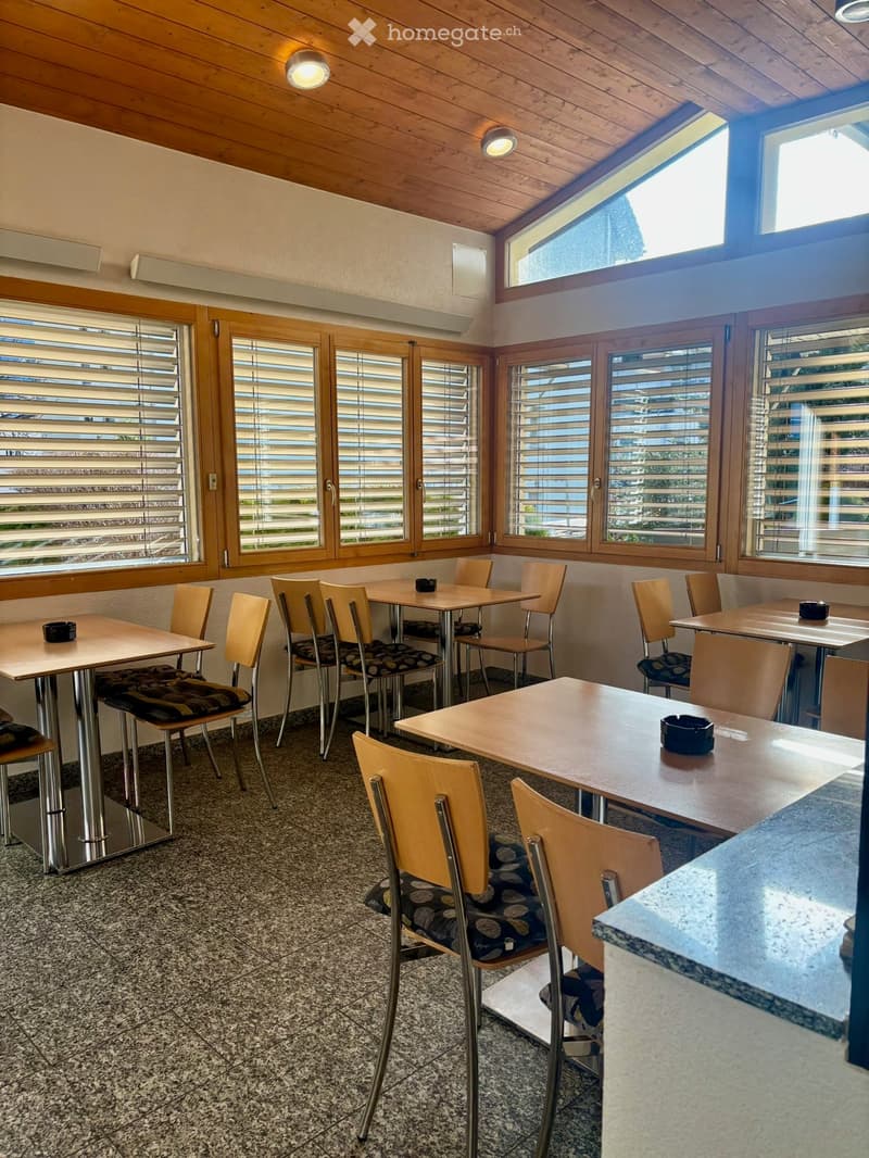 Brightly lit dining area with wooden tables and chairs, large windows with blinds, and a polished tiled floor.