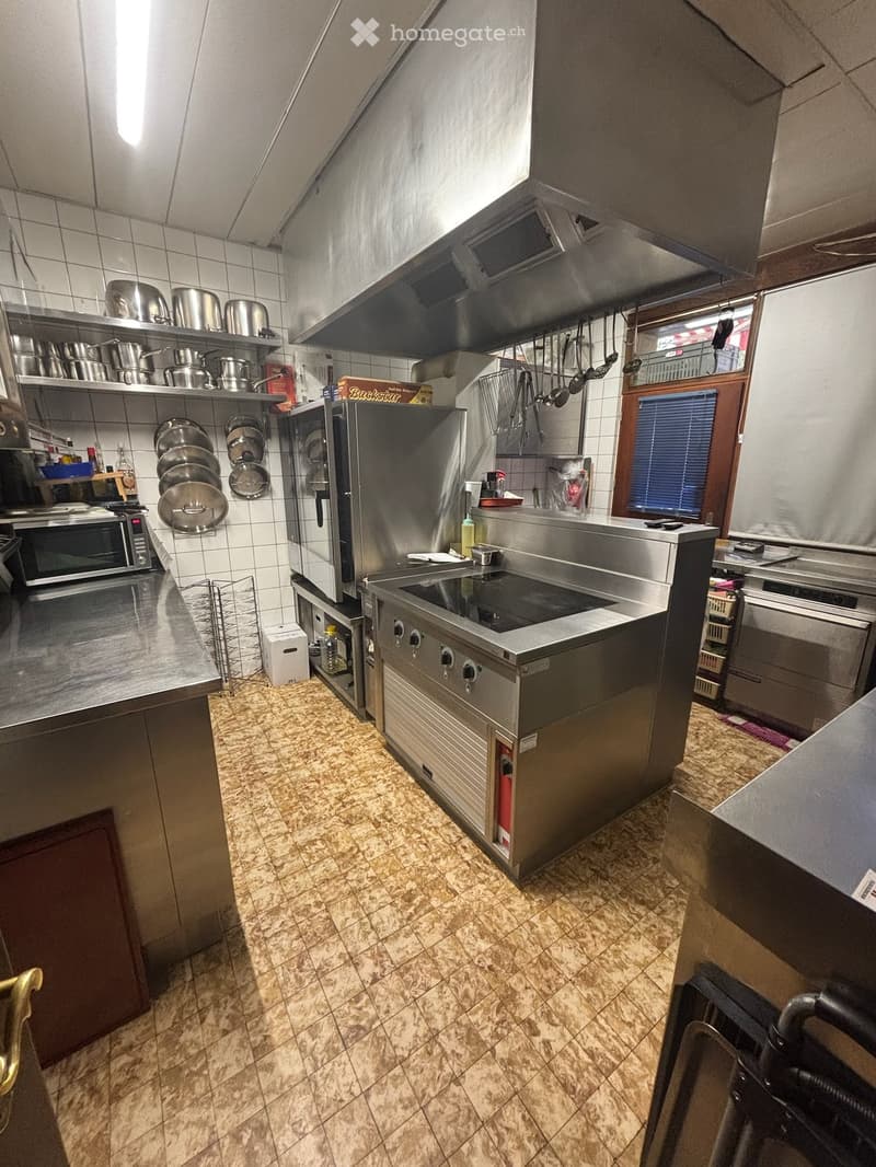 Commercial kitchen with stainless steel appliances, including a refrigerator, microwave, oven, and stove. Pots and pans are hanging and on shelves. The floor is tiled, and there is a window with blinds.