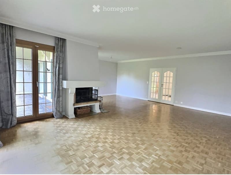 Empty living room with light grey walls, herringbone wooden flooring, fireplace, large glass doors, and double doors leading to another area.