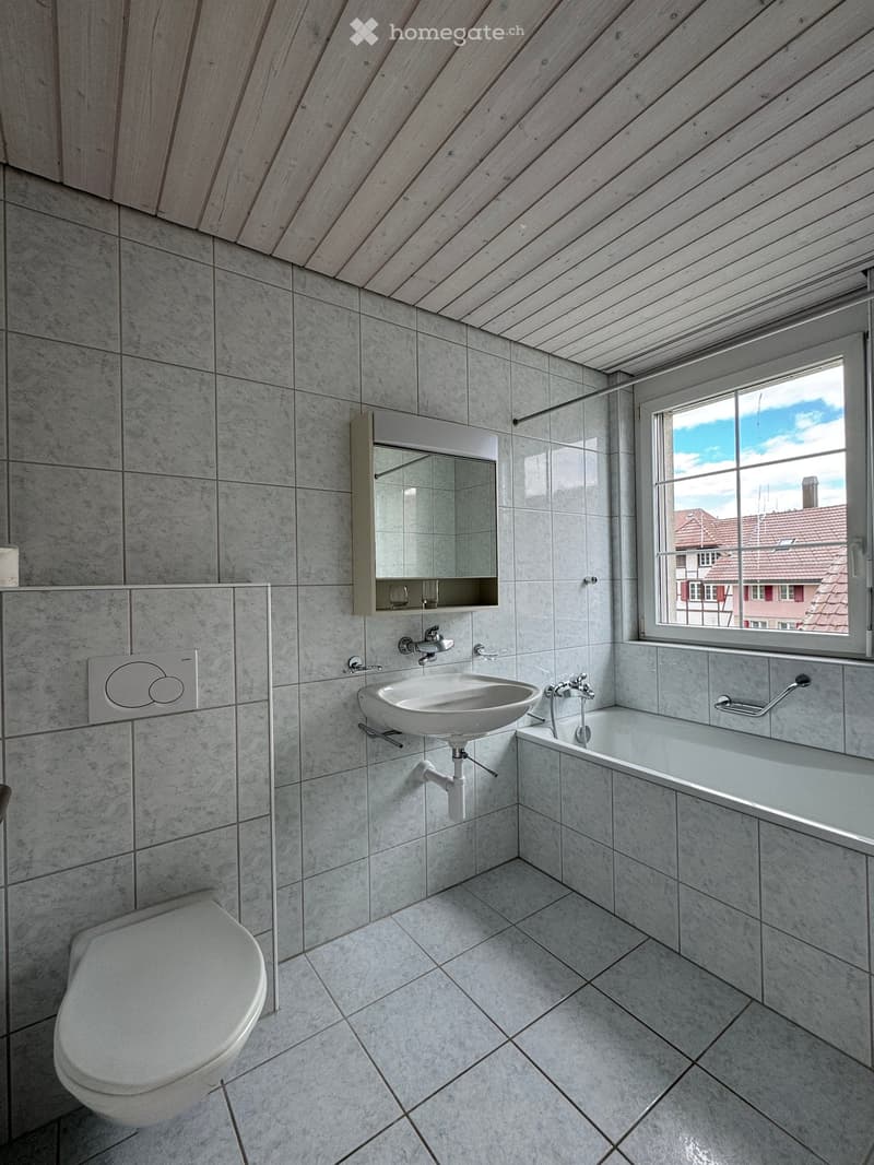 Modern bathroom with grey tiled walls and floor, featuring a toilet, sink, bathtub, and a mirror with a built-in shelf.