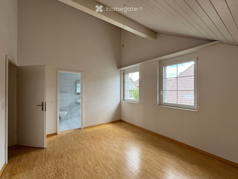 Empty room with wooden flooring, light-colored walls, and two windows. A door leads to a small bathroom with a toilet and sink.