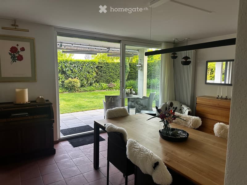 Bright living room with large sliding glass doors opening to a garden, wooden dining table with chairs, sofa, flower vase, and decorative items.