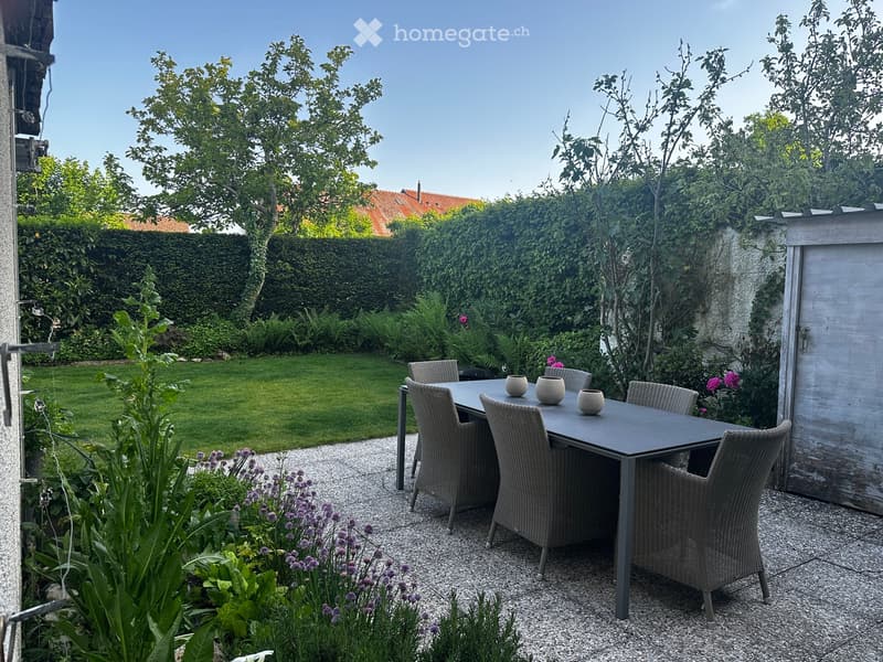 Garden with a patio area featuring a table and chairs, surrounded by greenery and a well-maintained lawn.