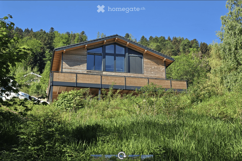 Modern two-story wooden house with large windows and a balcony, surrounded by lush greenery and trees.
