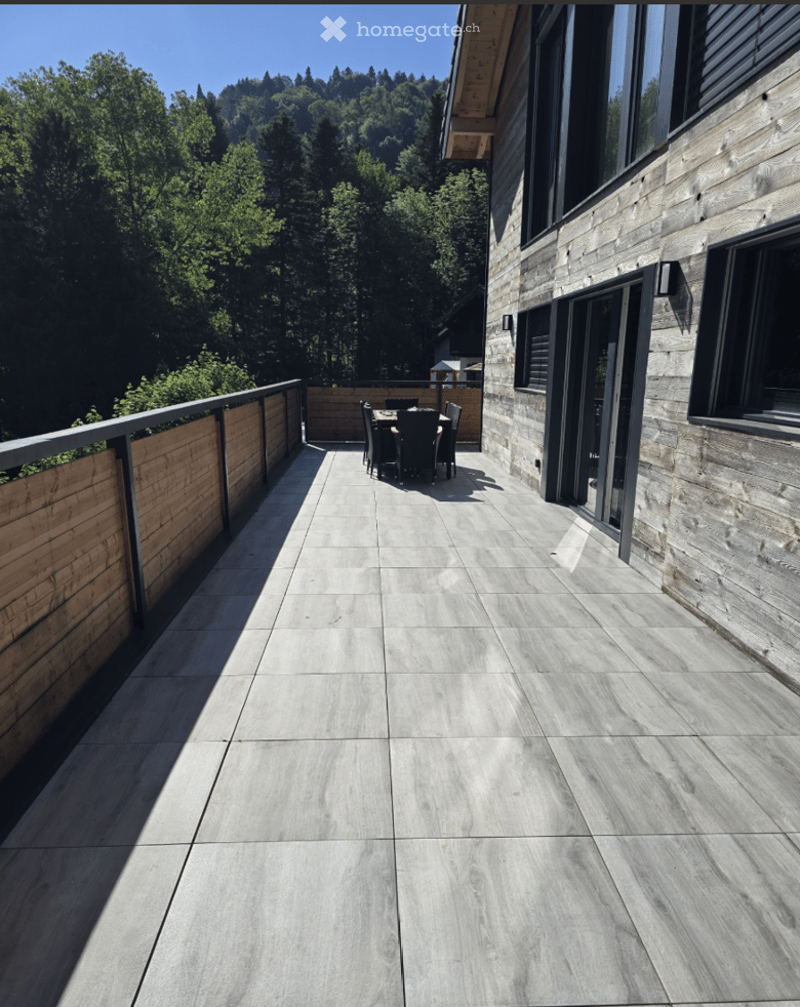 Modern balcony with grey tiled flooring, wooden railing, and a small dining set in the background, overlooking a lush green forest.