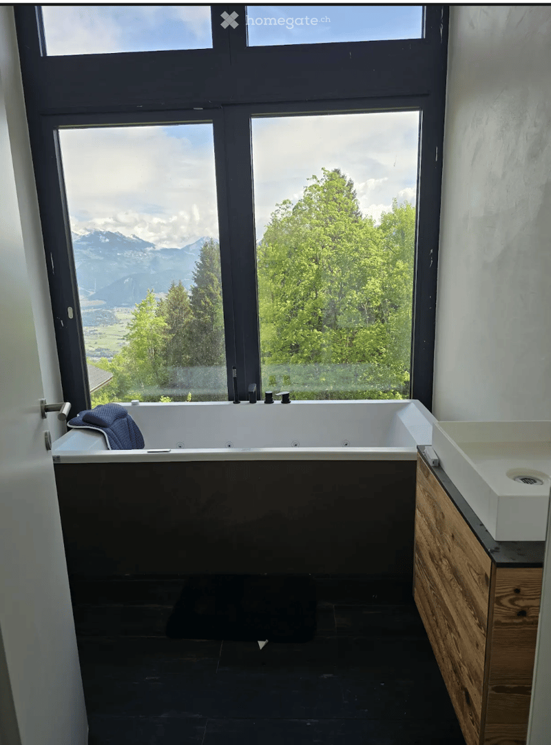 Modern bathroom with large windows offering a scenic mountain view, featuring a bathtub, sink, and a towel draped over the edge.