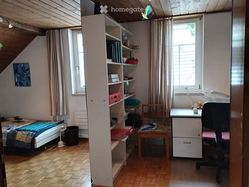 Cozy attic bedroom with wooden floors, slanted ceilings, and large windows. Features a bed, desk, bookshelf, and a small chair.