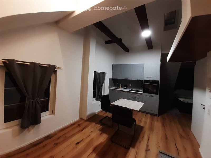 A modern, open-plan living and dining area with wooden flooring, grey kitchen cabinets, a dining table with chairs, and a window with dark curtains.
