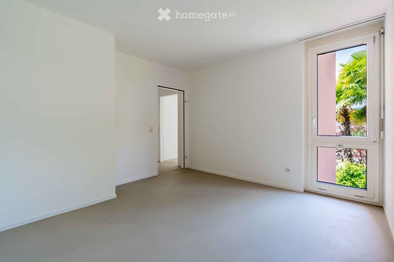 Empty room with white walls, grey carpeted floor, large window with blinds, and an open doorway leading to another space.