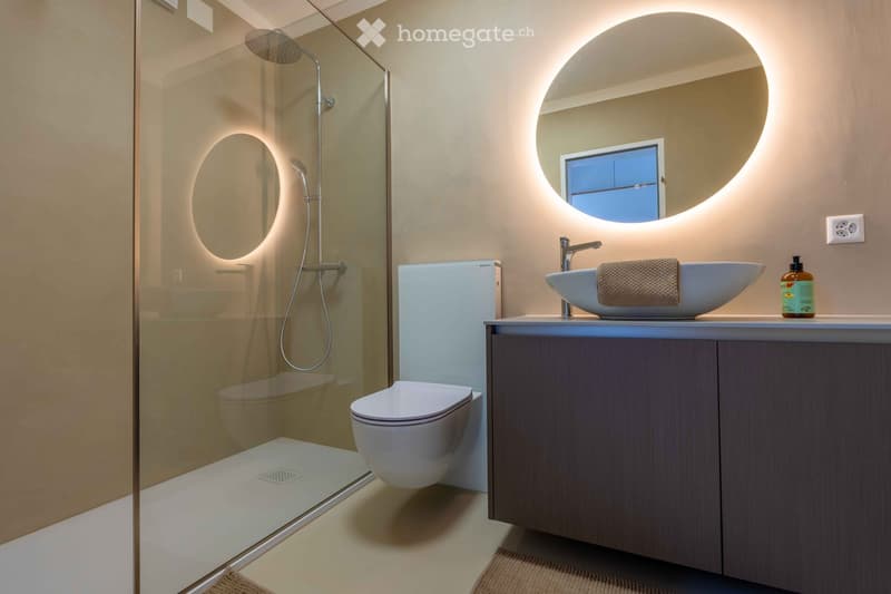 Modern bathroom with a glass-enclosed shower, a floating toilet, a sleek sink with a modern faucet, and a round illuminated mirror above the sink.