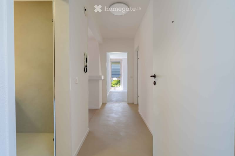 A clean, modern hallway with white walls, a smooth tiled floor, and a round ceiling light. The hallway features a door on the left and a window at the end, allowing natural light to enter.