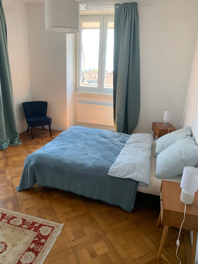 Cozy bedroom with wooden flooring, a large window with blue curtains, a neatly made bed with light blue bedding, a wooden nightstand with a lamp, a small wooden chair, and a patterned rug.