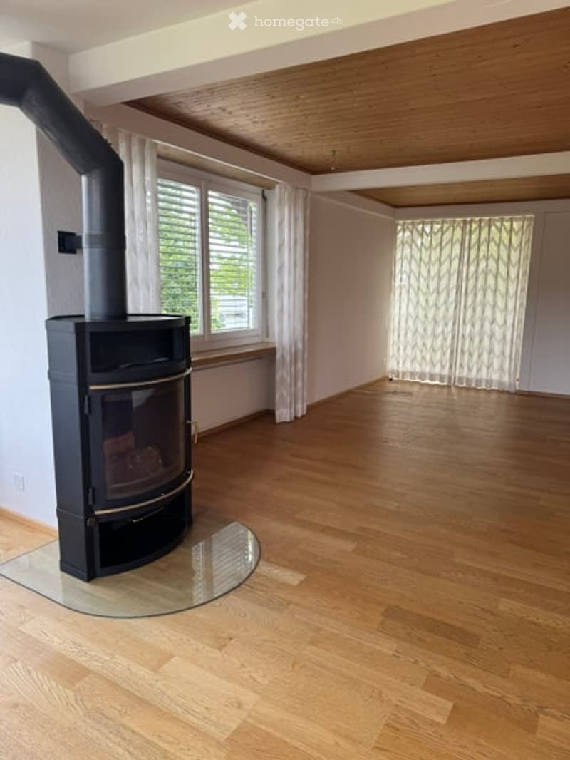 Empty room with wooden flooring, white walls, large windows with white curtains, and a black wood-burning stove with a chimney.