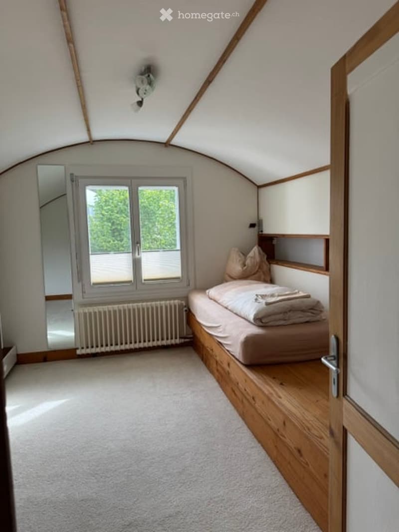 Cozy bedroom with a built-in wooden bed, pillows, blanket, and a large window offering a view of greenery outside.