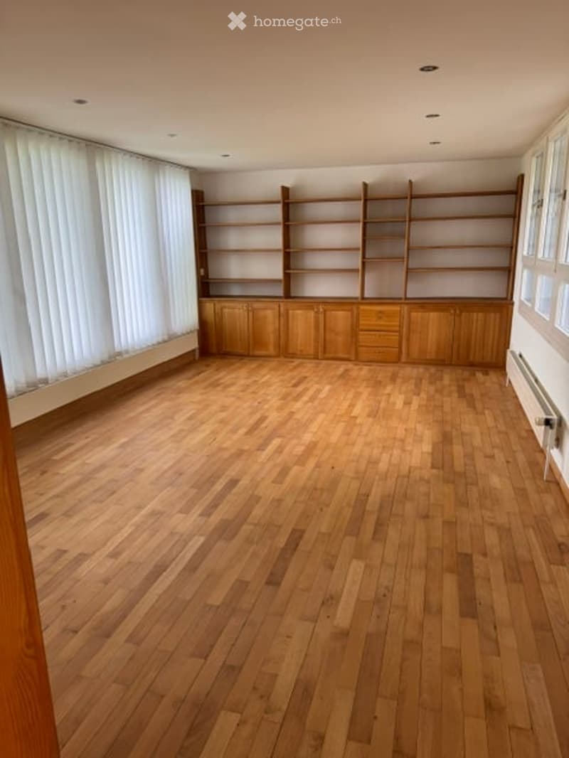 Empty room with wooden flooring, built-in wooden shelving unit, large windows with white blinds, and a radiator on the wall.