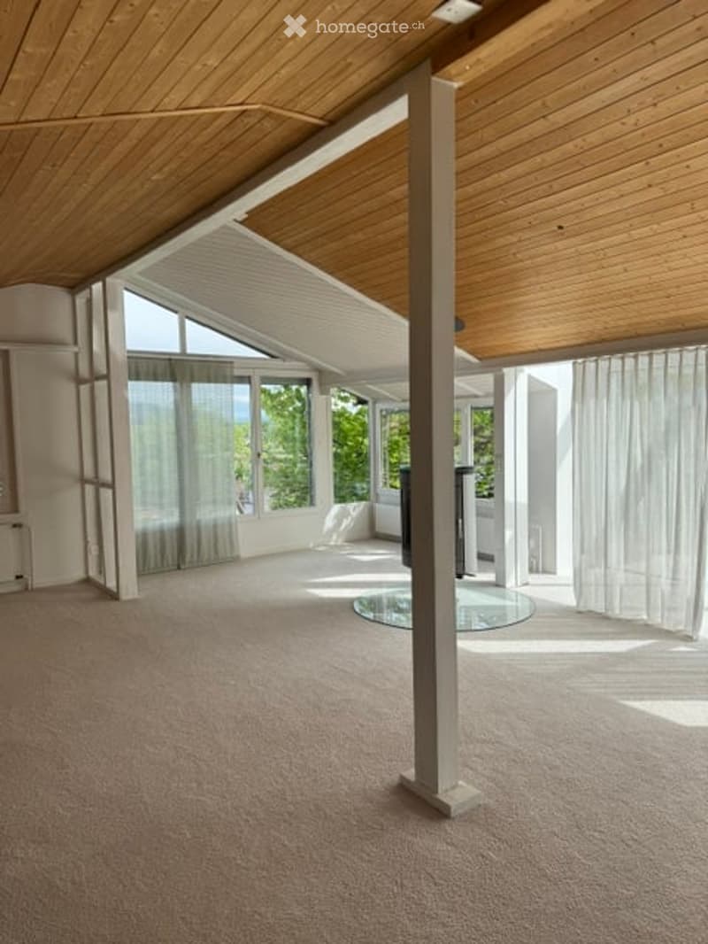 Bright, open loft-style room with wooden ceiling beams, large windows, and a central column. The space is completely empty with no furniture or equipment.