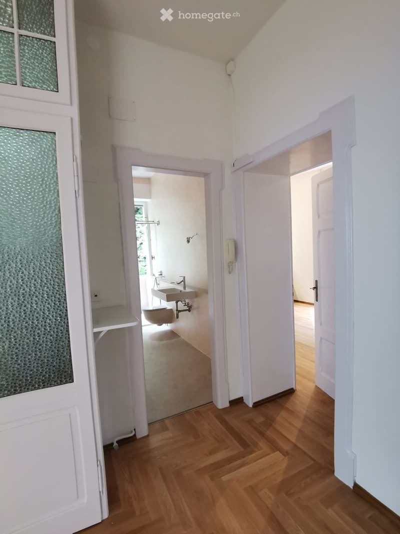 A narrow hallway with wooden flooring, white walls, and two open doors leading to adjacent rooms. One door reveals a modern bathroom with a sink, toilet, and a window, while the other leads to an unfurnished room with light-colored walls.