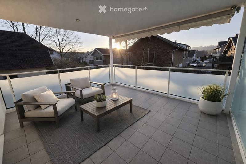 Balcony with two chairs, a small table, a potted plant, and a lantern, featuring a tiled floor and a view of a residential neighborhood at sunset.