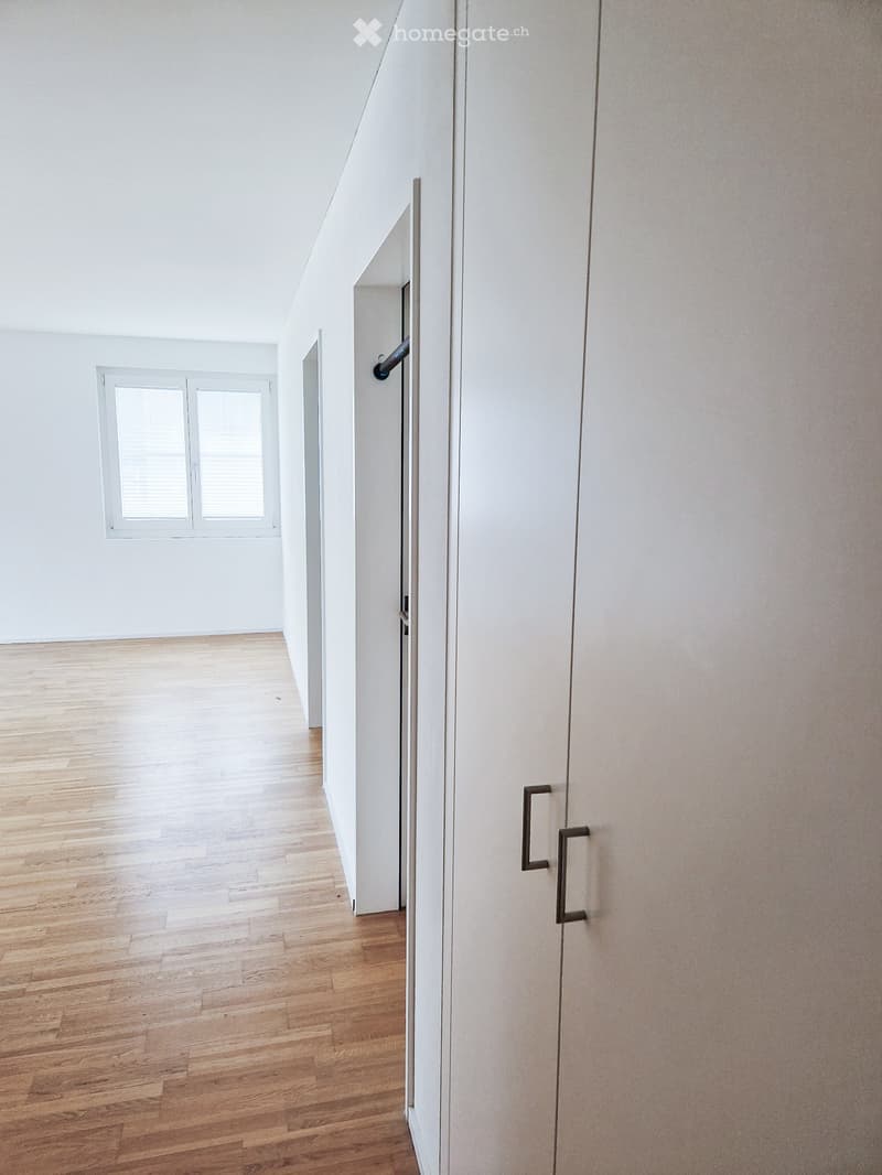 Empty hallway with wooden flooring, white walls, and a window with blinds. No furniture or equipment present.