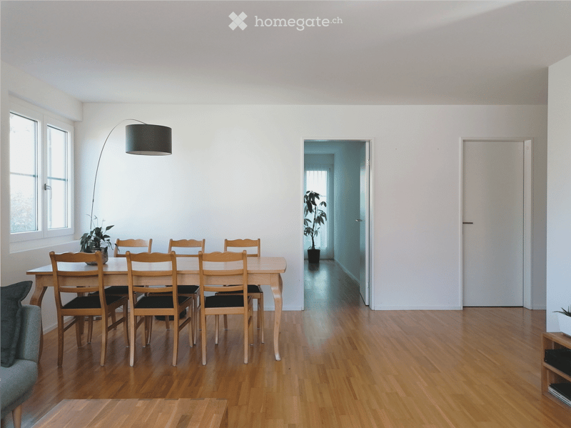 Bright dining area with wooden table, six chairs, floor lamp, and a small plant. Adjacent living space visible with a sofa and wooden flooring.