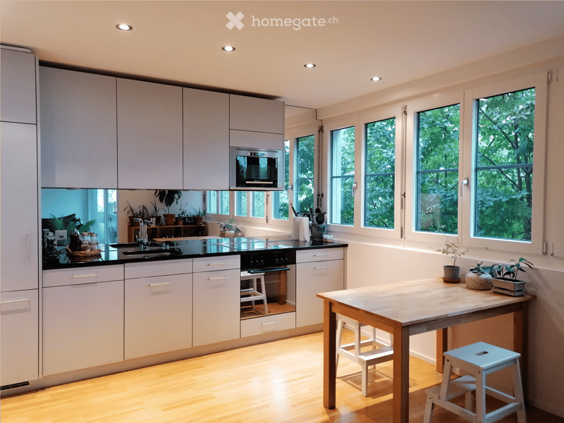 Modern kitchen with white cabinets, black countertops, and a central island table with stools. Features include a sink, stove, microwave, and several potted plants.