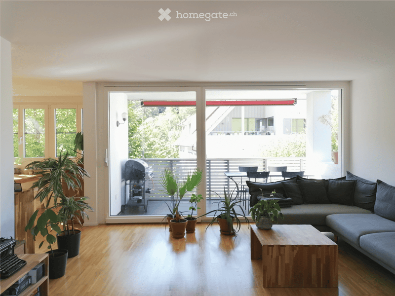 Bright living room with wooden flooring, large sliding glass doors leading to a balcony, sofa, coffee table, potted plants, and a small table with chairs on the balcony.