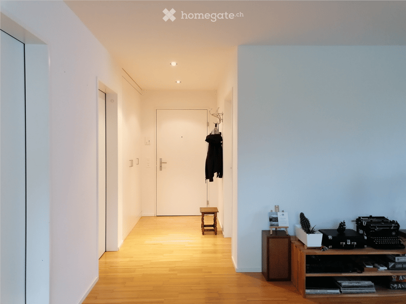 A modern hallway with wooden flooring, white walls, and several pieces of furniture including a small wooden stool, a cabinet, a shelf with a typewriter, a suitcase, and a potted plant. A coat rack with a jacket is visible, and a mirror is mounted on the