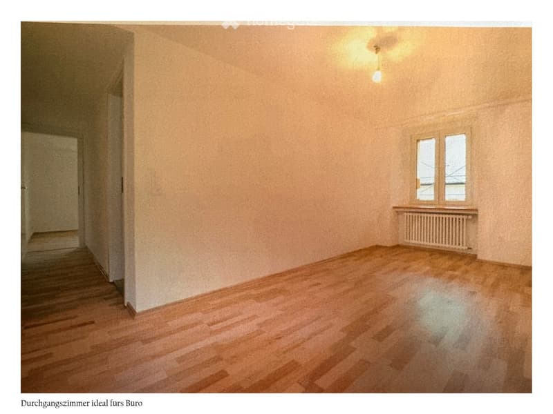 Empty room with wooden flooring, beige walls, and a window with a radiator below it. The room is unfurnished and has a doorway leading to another space.
