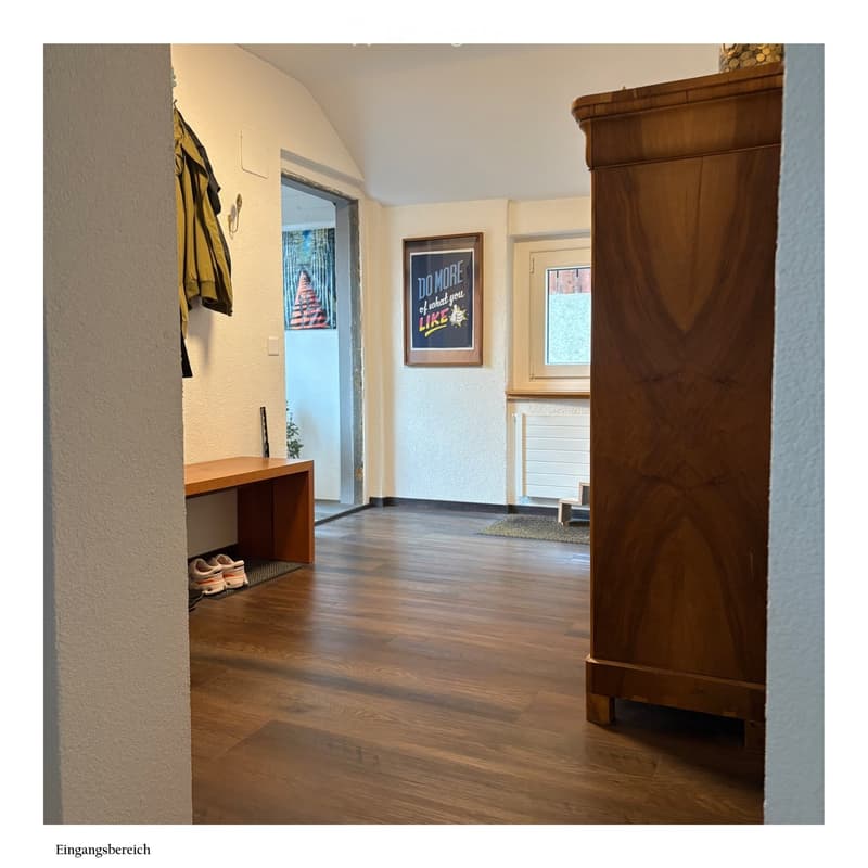 Spacious hallway with wooden flooring, a coat rack with jackets, a small bench, a framed motivational poster, and a window with a radiator below it.