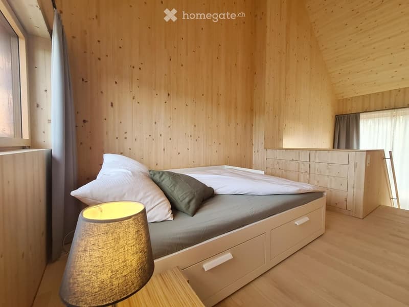 Single bed with drawers, wooden walls, white bedspread, light colored floor, bedside lamp, curtains