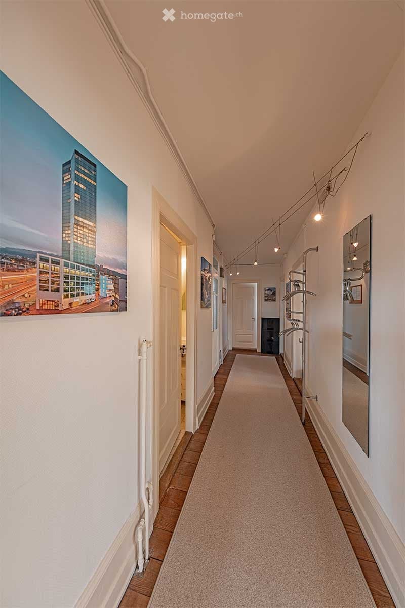 Long hallway, beige carpet, picture on the wall, radiator, and mirror