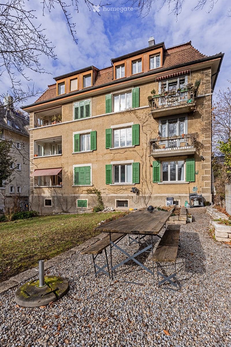 A three-story residential building with beige stone facade, green shutters, multiple balconies, and a wooden picnic table in the front yard.