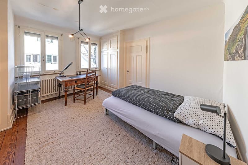 wooden desk and wooden chair, small double bed, lamp on the side, radiator, large windows