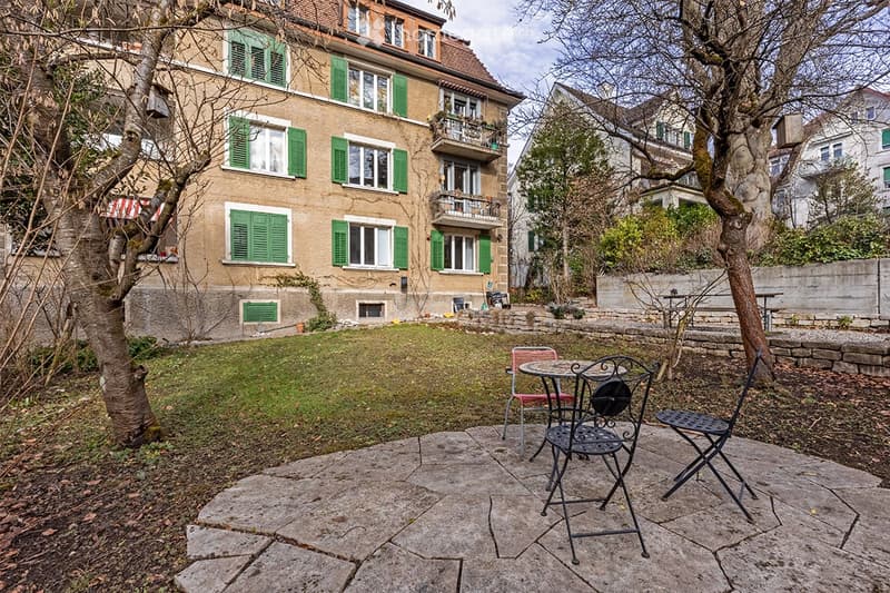 Garden area with a stone patio, outdoor furniture, and a multi-story building with green shutters in the background.